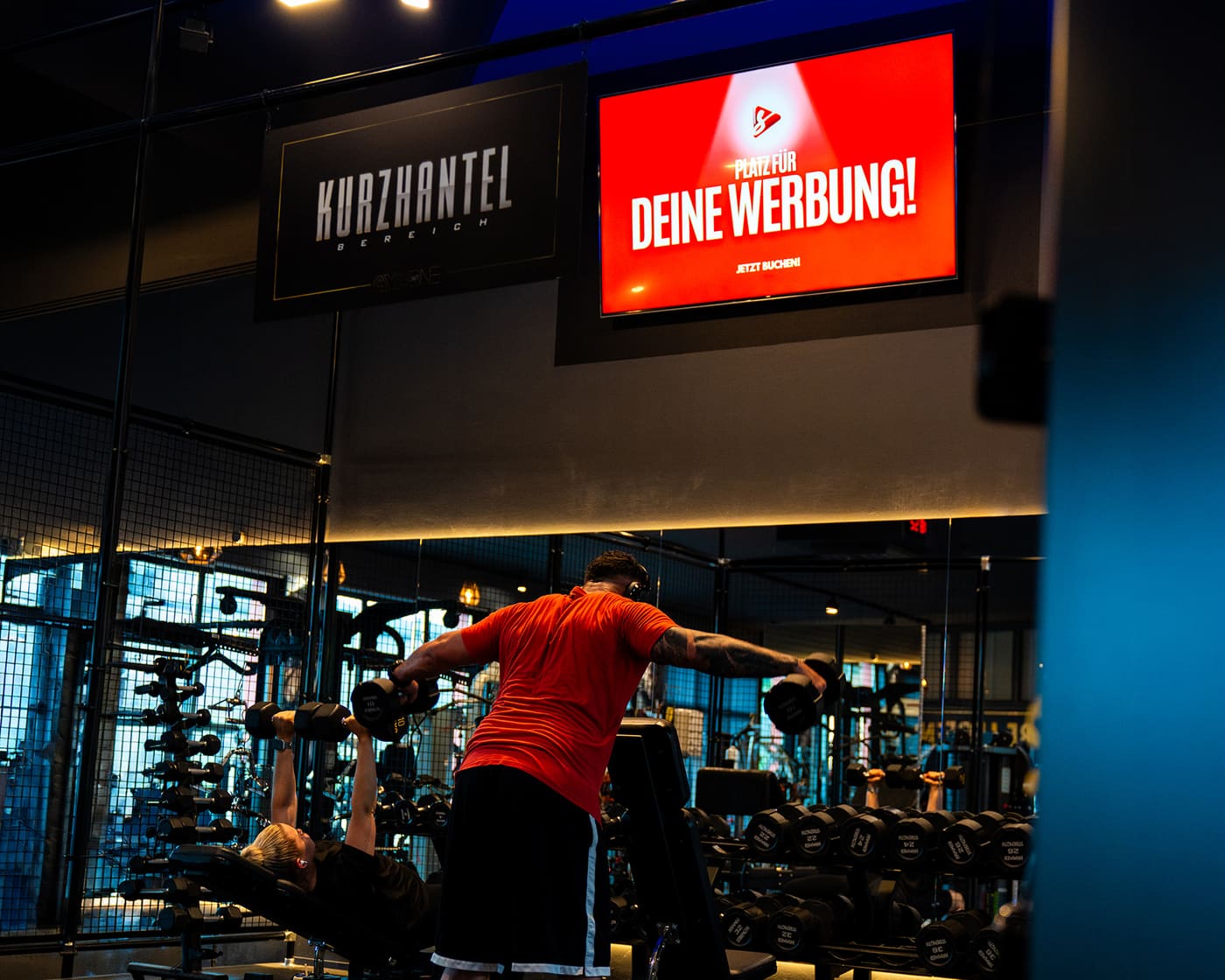Bildschirm mit der Anzeige Deine Werbung im Kurzhantelbereich des Fitnessstudios GymOne