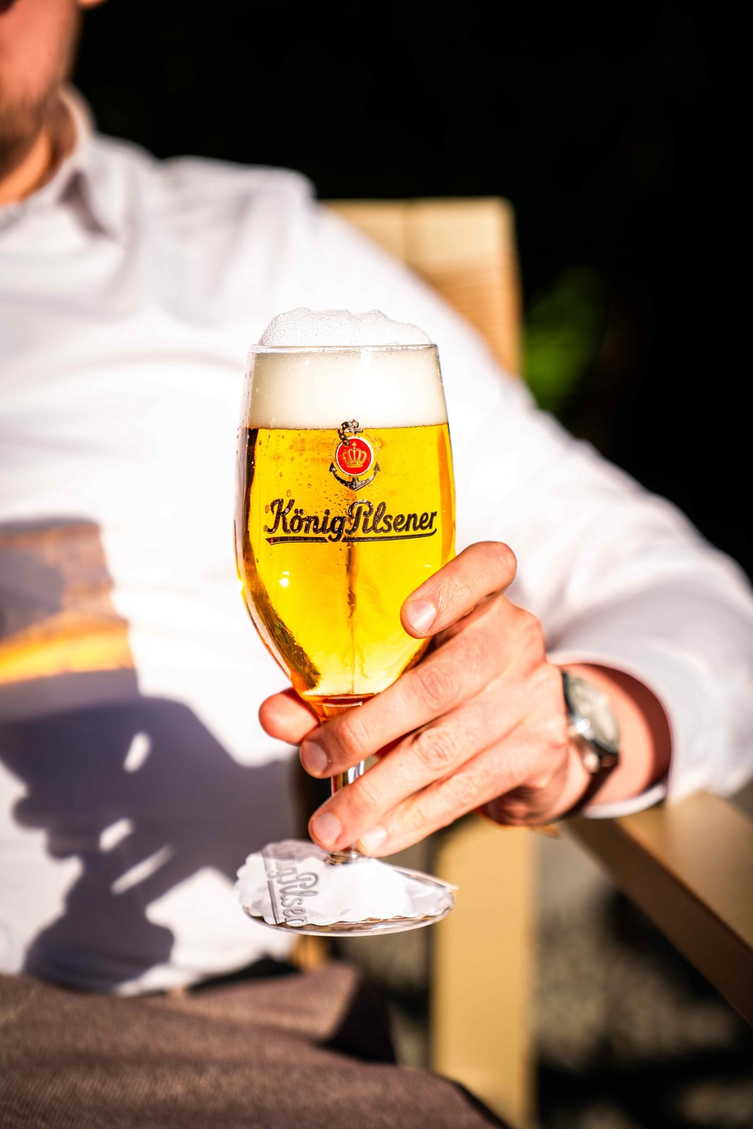 Hand hält ein gekühltes Glas König Pilsener im Gegenlicht der Abendsonne